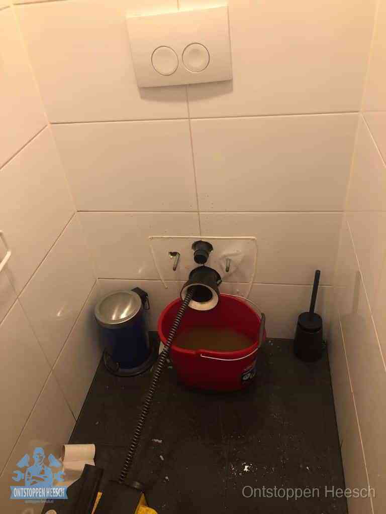 Een ontstopte wand-wc met verwijderde toiletpot waar een ontstoppingsveer in de wanduitgang verdwijnt, het achterhalen waar de verstopping zit in Heesch