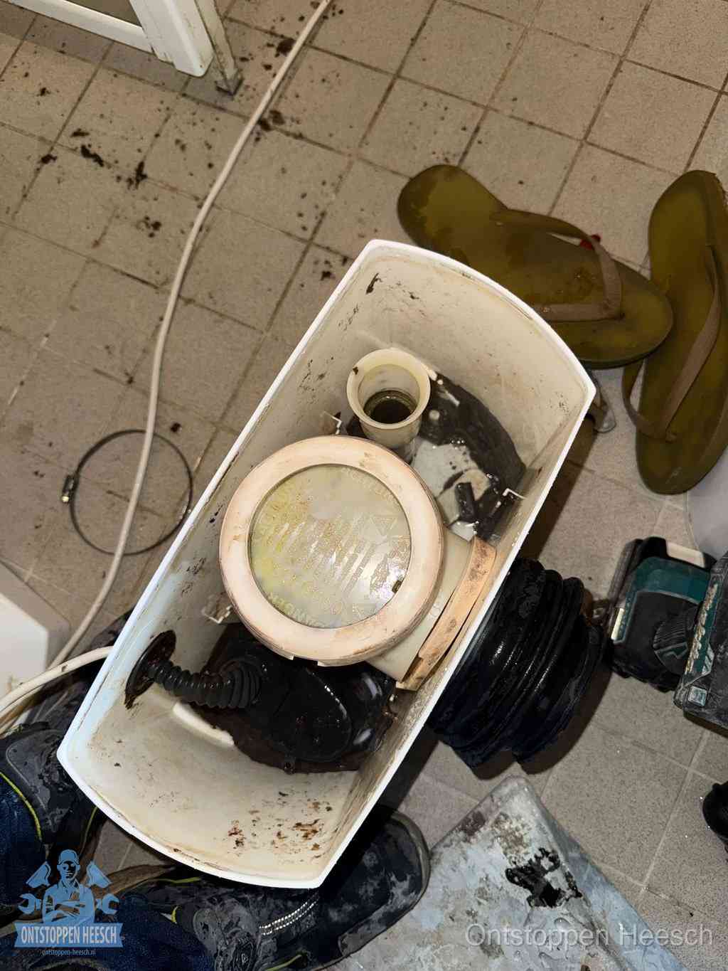Een gedemonteerde toiletpot op de vloer met ontstoppingsveer en gereedschap ernaast, het verwijderen van een vastzittend voorwerp uit de wc in Heesch